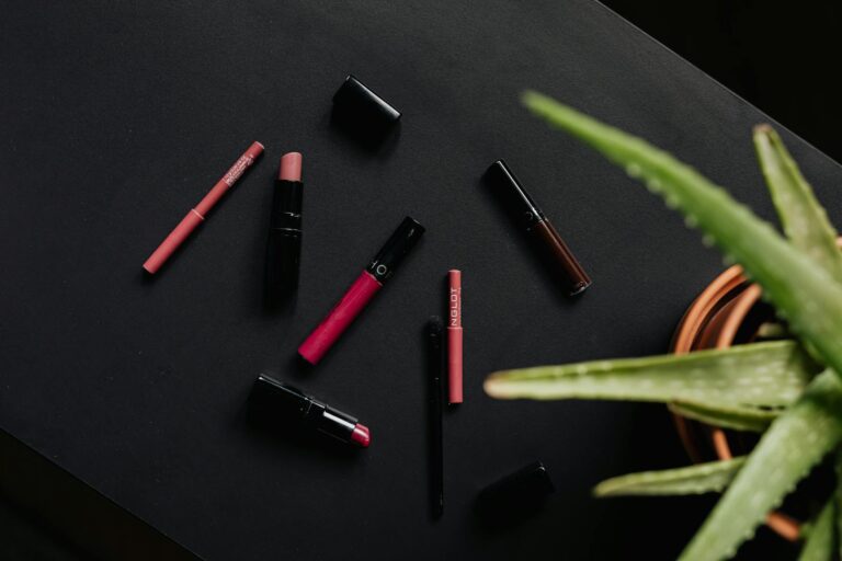 Come proteggere i capelli 1 Overhead view of an assortment of lipsticks and makeup products complemented by an aloe plant, on a black background.