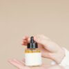 Close-up of hands holding a glass dropper bottle for skincare on beige background.